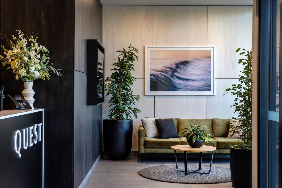 Level grey tiled reception with a wide, clear path. Olive green sofa seating, round coffee table on a rug, and large potted plants. High-contrast 'QUEST' signage on a dark reception desk.