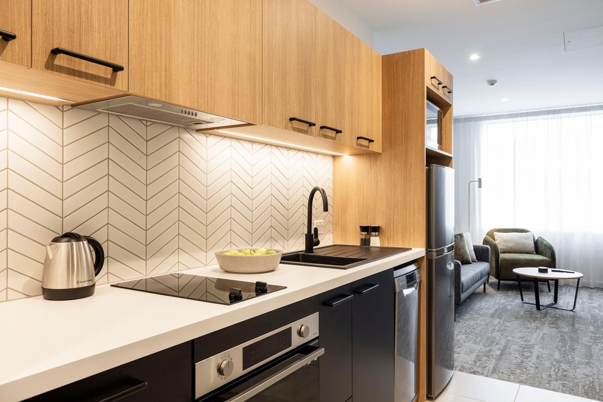 Level tiled kitchen floor transitions to a grey carpeted living area with a sofa, armchair, and coffee table. Features white benchtops and bright recessed ceiling and under-cabinet lighting.
