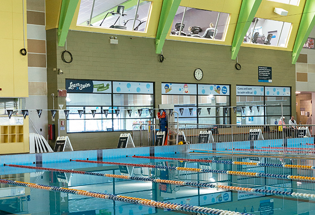 Indoor pool with lane ropes and silver handrails leading into a ramped entry. Large windows display 'Learn to Swim' signs below bright green structural beams and grey tiled pool deck.