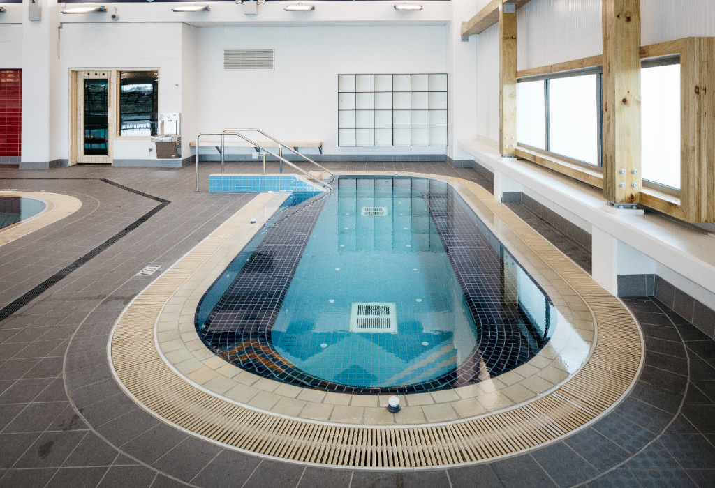 Long oval pool with blue tiles, ramp entry and stainless steel handrails. Level grey tiled floor. Bright indoor lighting with timber window frames and white lockers in the background.