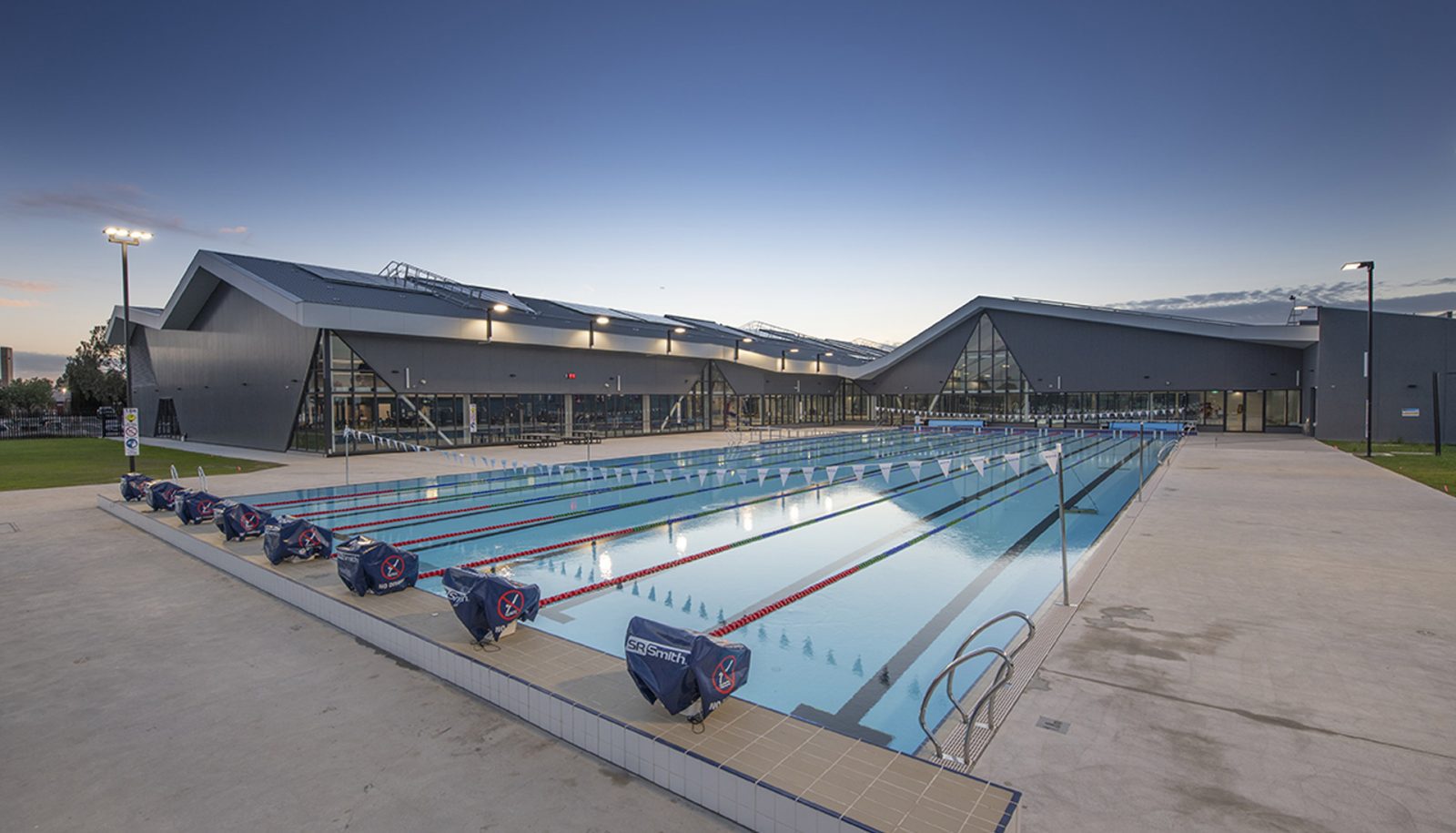Outdoor lap pool with wide level concrete surrounds and silver handrails. Dark grey building features glass windows and level entry points. Tall poles provide outdoor lighting.
