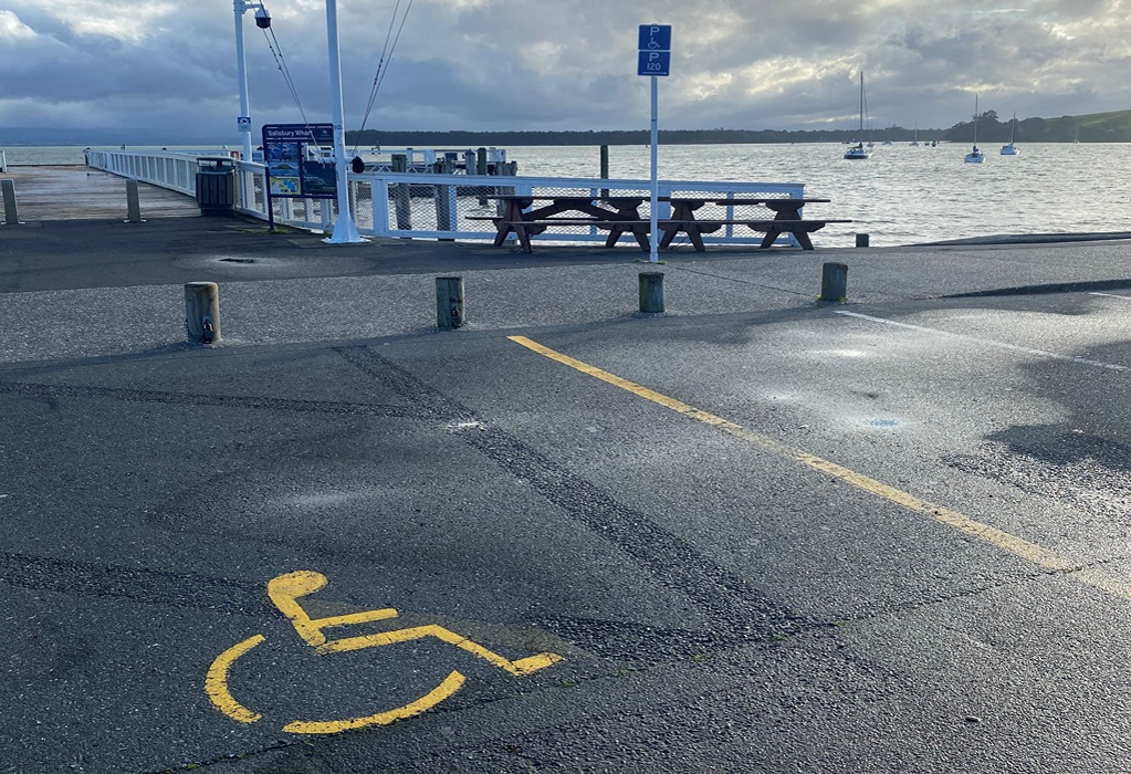Level asphalt parking space with yellow wheelchair symbol and blue accessible parking sign. Wooden bollards border a flat wharf with picnic tables and white railings.