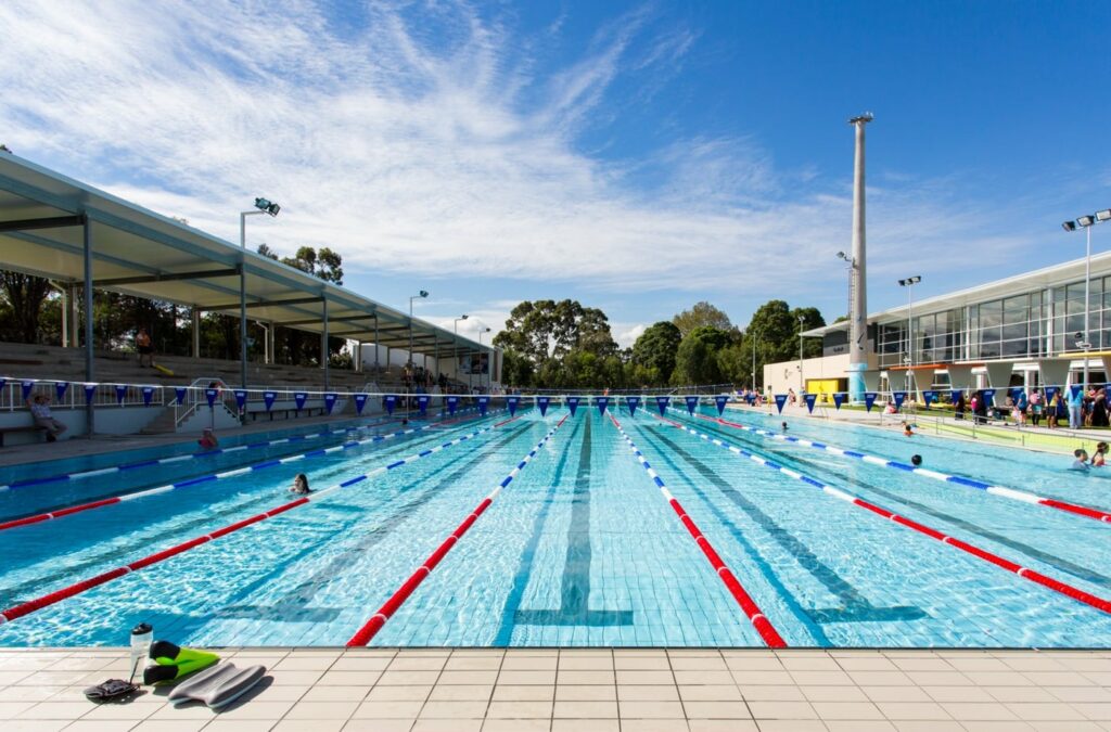 Outdoor 8-lane pool with wide, level light-coloured tiled surrounds. Covered tiered concrete seating on the left. Wide, step-free access paths lead around the pool deck.