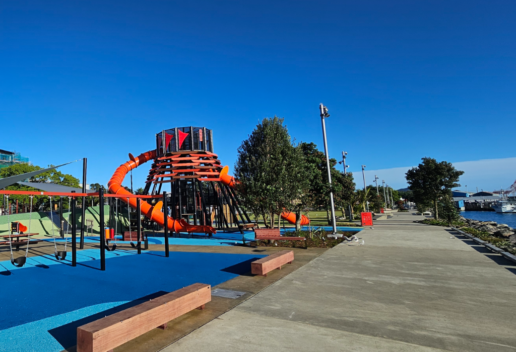 Wide level concrete path beside blue rubber safety surfacing. Timber block benches and benches with backrests. Tall grey lamp posts. Large orange climbing tower. Clear, level access throughout.