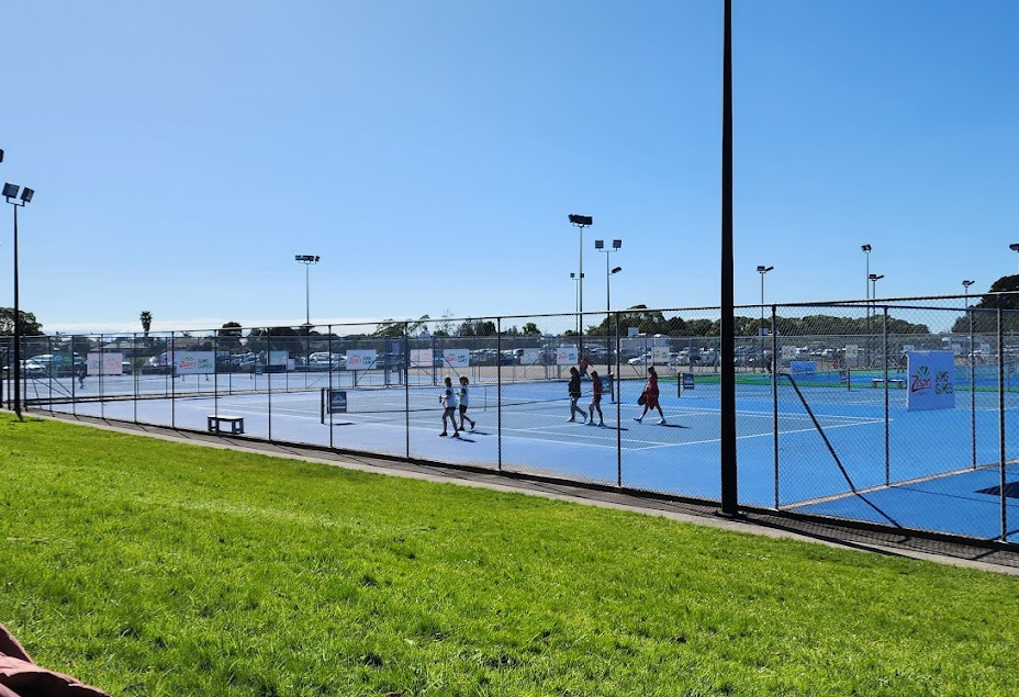 Level blue hardcourt tennis courts enclosed by chain-link fencing. Grassy area in foreground. Tall floodlight poles, white banners and a bench. Parking area visible in the background.