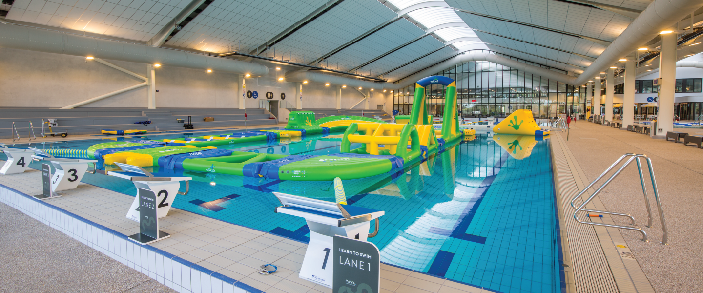 Indoor pool with green and yellow inflatable course. Numbered starting blocks, stainless steel handrail at entry, non-slip grey floor, and blue accessibility signage on the far wall.