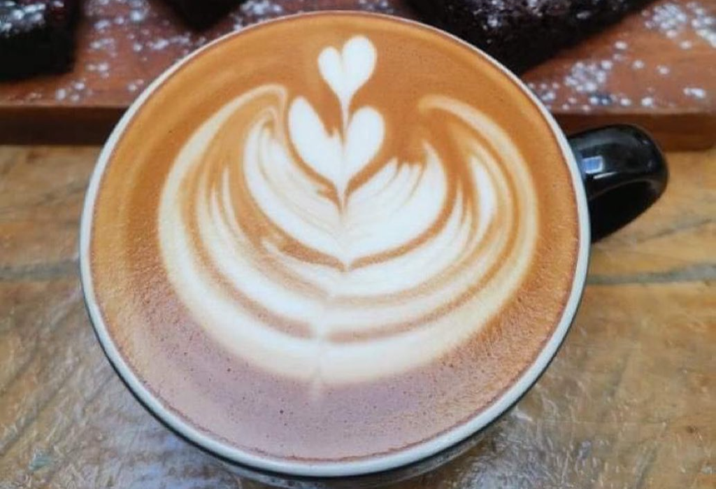 White latte art in a rosette and double heart pattern on light brown coffee in a black mug. The mug sits on a textured light brown tabletop.