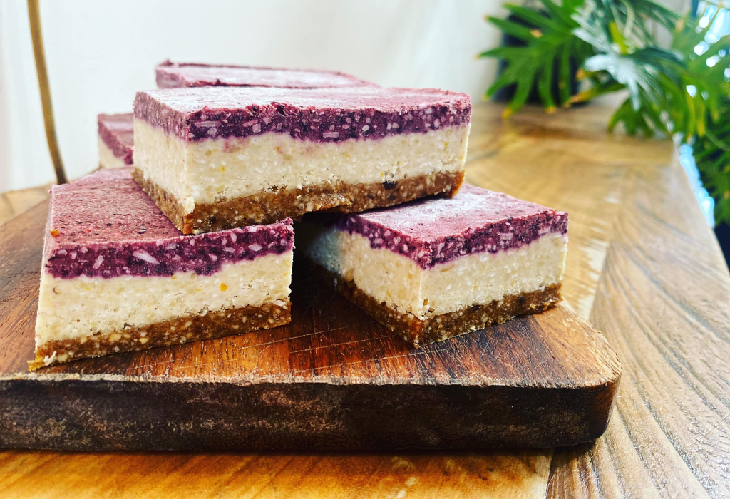 Three rectangular layered slices on a dark timber board. Crumbly brown base, thick white centre, and purple top with white flecks. Rustic wooden surface with green foliage behind.