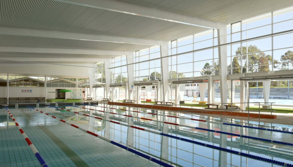 Indoor lap pool with blue tiled floor and lane ropes. Floor-to-ceiling windows. Wide level concrete deck with metal handrails along a ramped entry. Timber benches and clear floor surfaces.