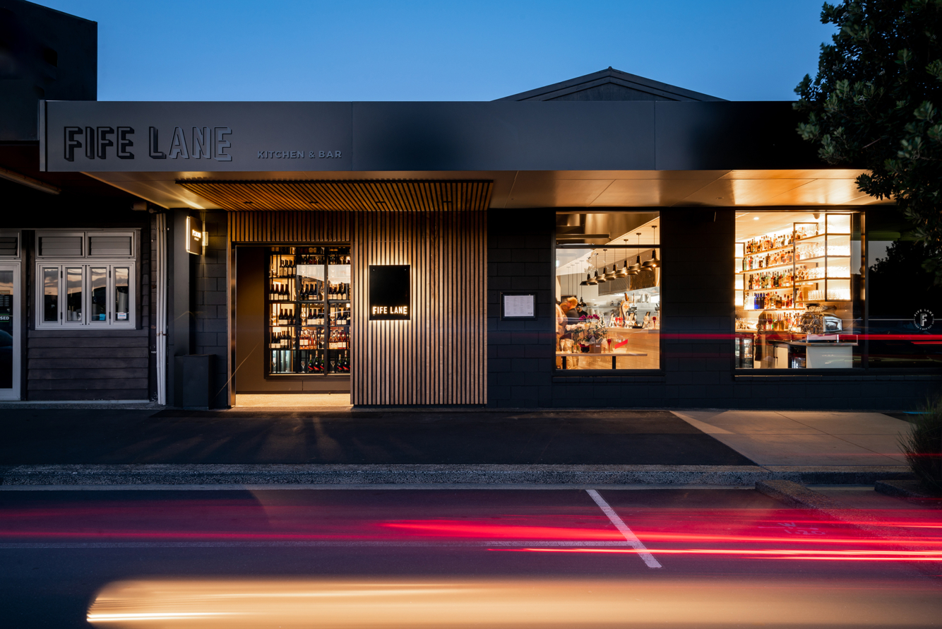 Level asphalt pavement leads to a wide, recessed entrance under a timber-slatted ceiling. Large glass windows show a lit interior. Dark brick exterior features illuminated "FIFE LANE" signage.