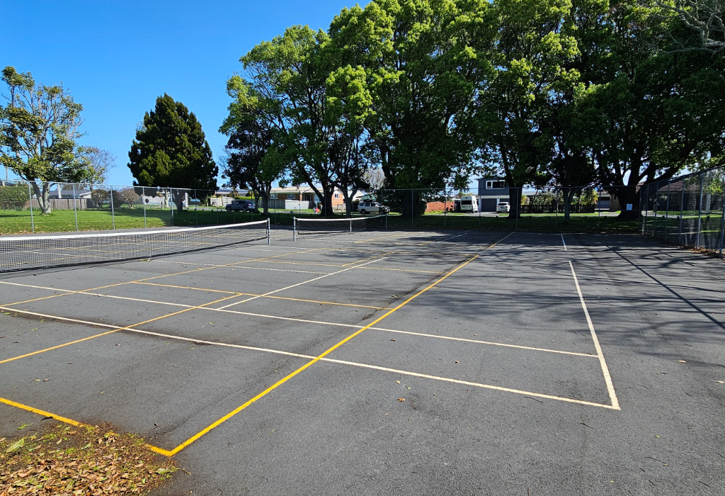 Level asphalt tennis court with yellow and white markings and a central net. Surrounded by a chain-link fence and large green trees. The flat ground surface is level with no visible stairs.