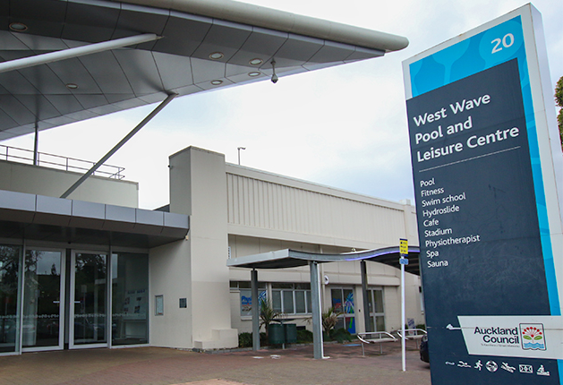 Level paved entrance with glass sliding doors under a grey canopy. A tall blue sign lists West Wave Pool and Leisure Centre facilities. Metal benches sit near a secondary covered walkway.
