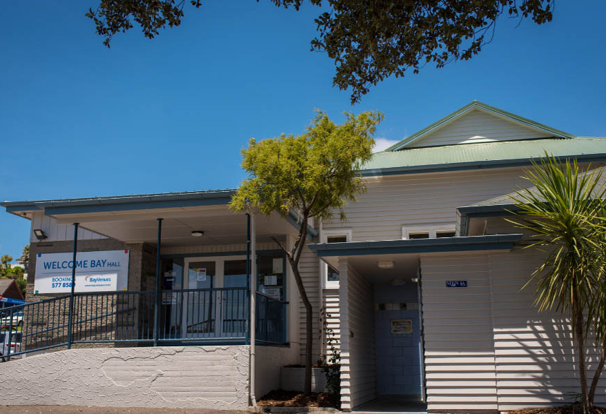 Welcome Bay Hall entrance with a concrete ramp and blue metal handrails leading to double glass doors. Large white and blue signage displays venue name and booking phone number.