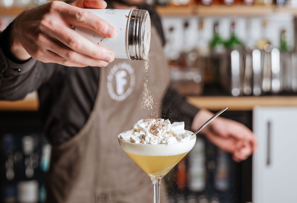 Person in a brown apron sprinkles powder from a shaker onto a cocktail with foam and meringue in a stemmed glass. Blurred wooden shelves with bottles are in the background.