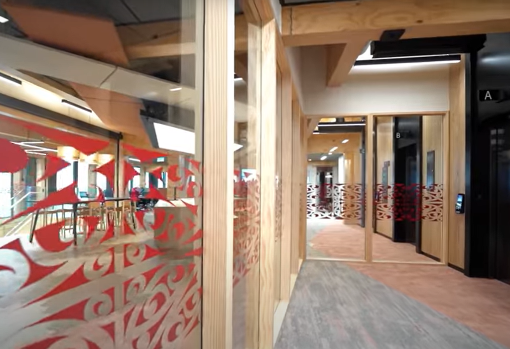 Bright, wide level hallway with grey and tan carpet. Timber-framed glass walls have red patterns. Path leads to black lifts marked A and B with a wall-mounted access panel.