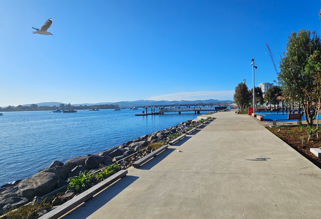 Wide, level concrete walkway with low timber edge beams and timber benches. A playground with blue rubber surfacing is on the right. Tall metal light poles are positioned along the path.