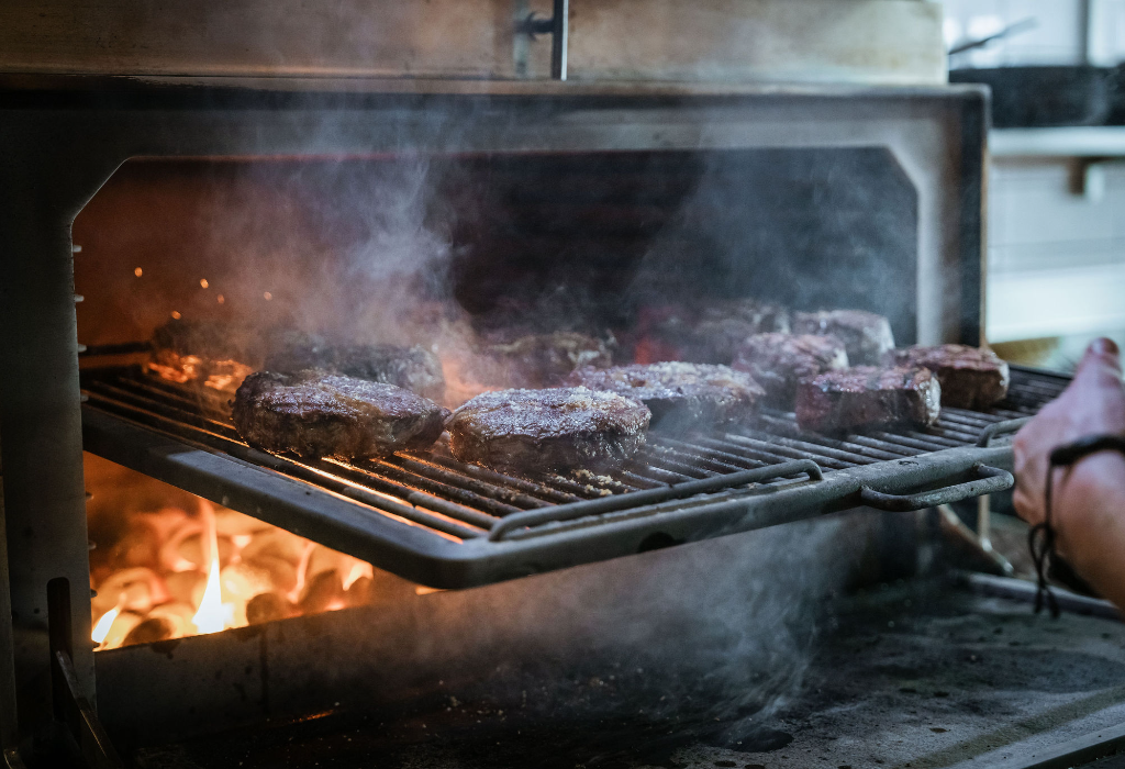 Metal grill rack with several thick steaks pulled from a charcoal oven. Bright orange flames and embers glow below the rack. Thick white smoke rises within the metal compartment.