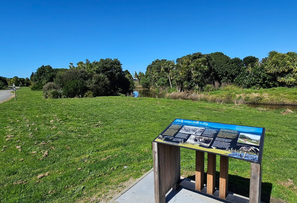 Tilted interpretive sign on a timber stand with a level concrete base. Surrounded by a flat mown grass field with level terrain. A gravel path is visible in the background.