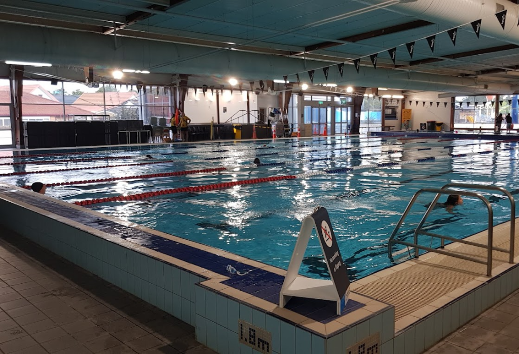 Indoor swimming pool with level tiled deck and metal handrail for access. Clear '1.8m' depth markers on edge. Wide, flat pathway leads to glass exit doors. Bright overhead lighting throughout.