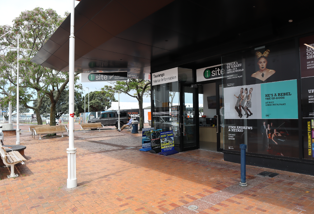 Level red brick paving leads to a visitor centre with a flat glass entrance. Timber benches are on the left. Portable signs sit on the flat ground under a large, dark overhanging canopy.