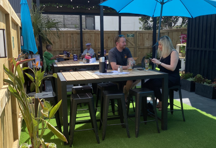 Level outdoor patio with green artificial turf. High timber-topped tables and black backless metal stools. Blue umbrellas provide shade and overhead string lights are beneath a dark canopy.