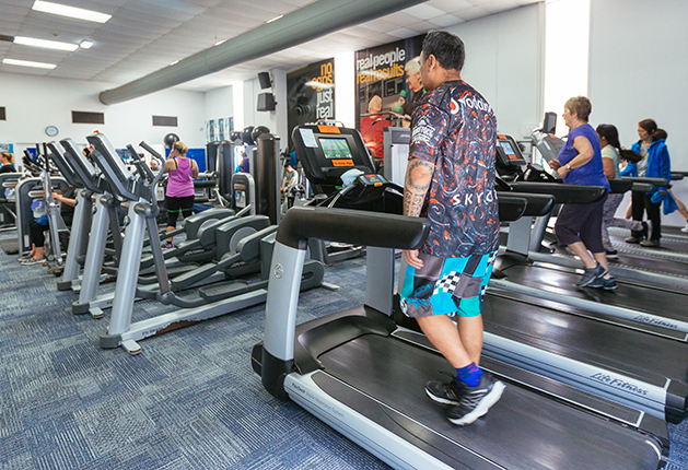 Level, blue-grey carpeted floor with wide pathways between rows of treadmills and ellipticals. Bright, even overhead lighting throughout the open-plan fitness centre.