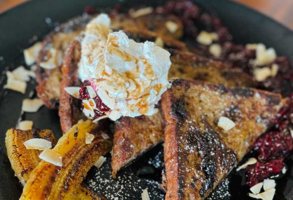 Golden-brown triangular French toast on a dark plate, topped with whipped cream, syrup, deep-red berry compote, toasted coconut flakes, caramelised banana slices, and a dusting of icing sugar.