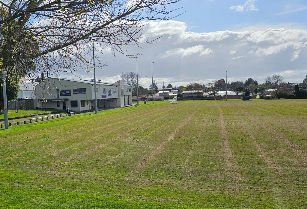 Grassy sports field with faint track lines. Two-storey light-coloured building with wooden bench seating. Level grass approach to building entrance. Tall silver light poles around the perimeter.