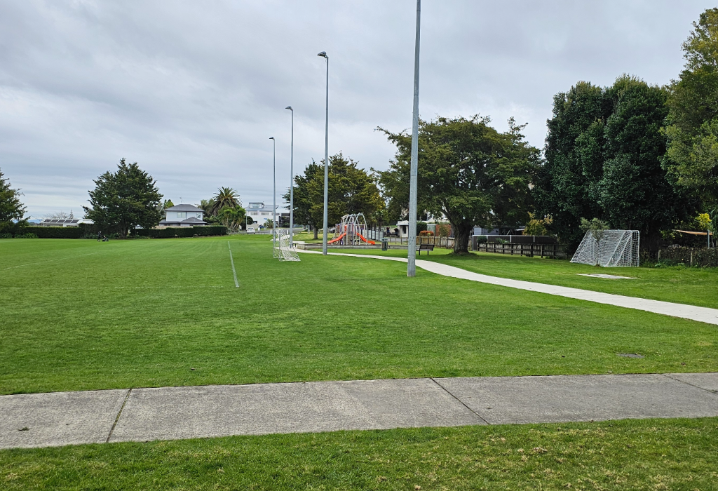 Wide, level concrete pathways wind across a flat grass park with netted soccer goals. A timber bench sits by the path near a playground. Tall grey light poles line the smooth paths.