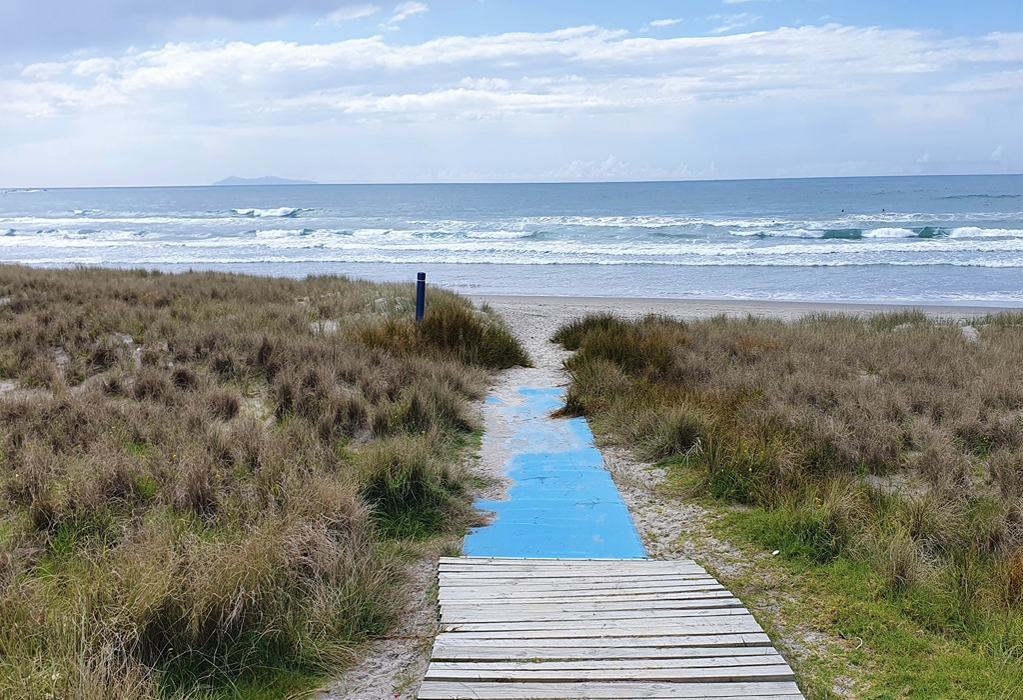 Wide, level slatted wooden boardwalk transitions to a bright blue accessibility mat path through sand dunes to the ocean, providing a flat, stable surface across the sand.