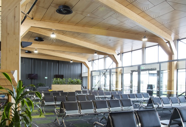 Level carpeted waiting area with rows of black chairs. Glass doors and windows provide flat access and natural light. High ceiling with timber beams and bright overhead lighting.