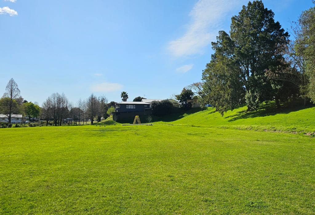Large, level green grass field with a grassy slope leading to trees. A yellow metal A-frame stands near a dark-coloured house in the distance. No paved pathways or seating are visible.