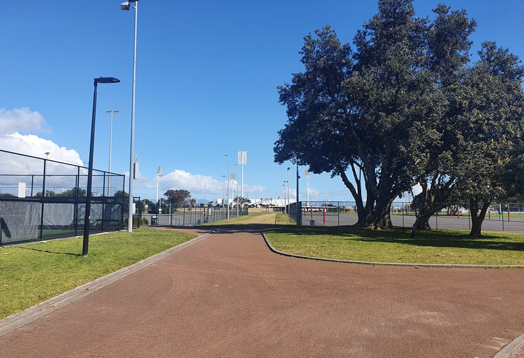 Level, wide reddish-brown paved path with concrete kerbs between grassy areas and chain-link fences. Tall light poles and trees line the route leading towards distant stadium light towers.
