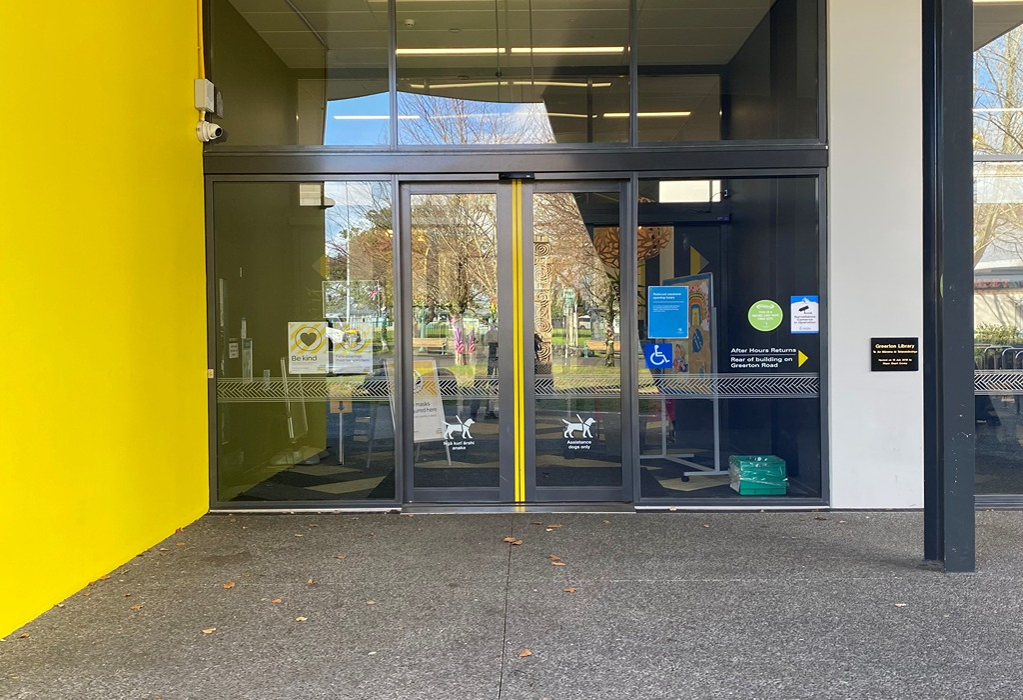 Level concrete entrance to Greerton Library with automatic glass sliding doors and yellow safety stripes. Signage includes a wheelchair symbol, assistance dog icons, and a returns arrow.