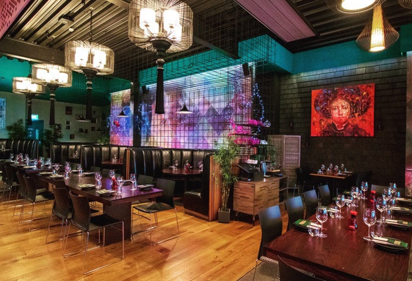 Level timber floor with wide pathways between dark wood tables, low-back black chairs, and leather booths. Dimly lit by birdcage chandeliers with purple and blue backlit feature walls.