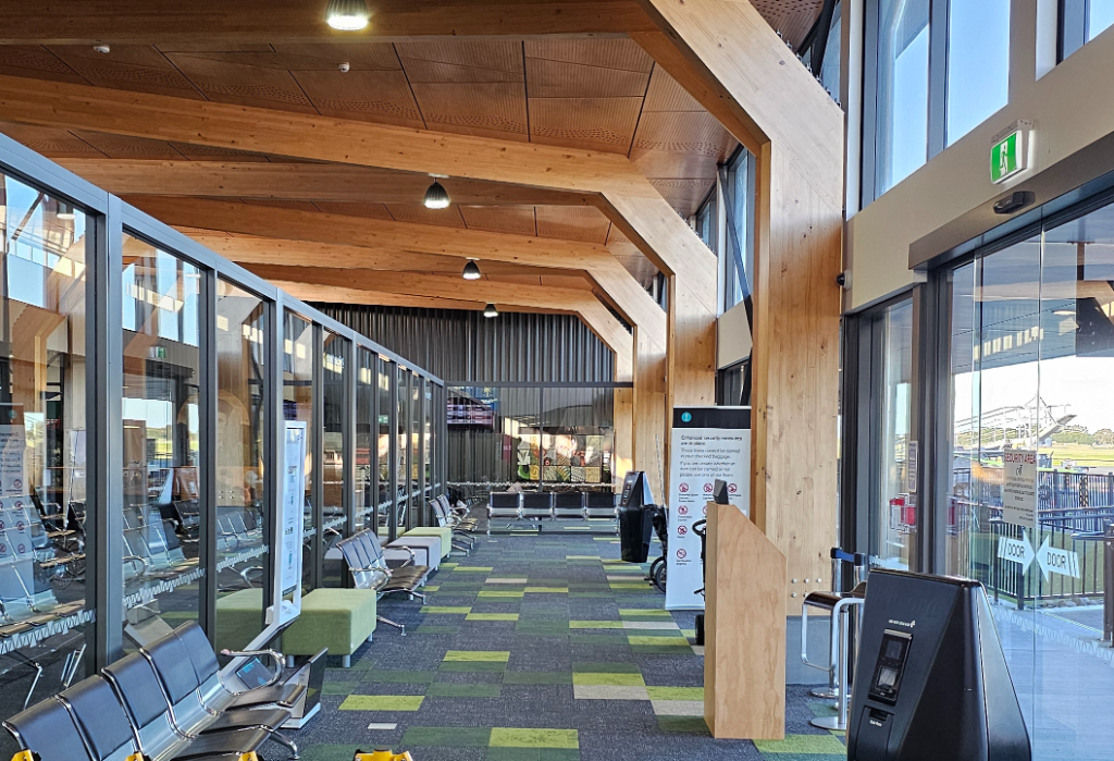 Wide level waiting area with green and grey patterned carpet. Rows of metal seating face glass walls. Level access to automatic glass doors on the right under high arched timber beams.