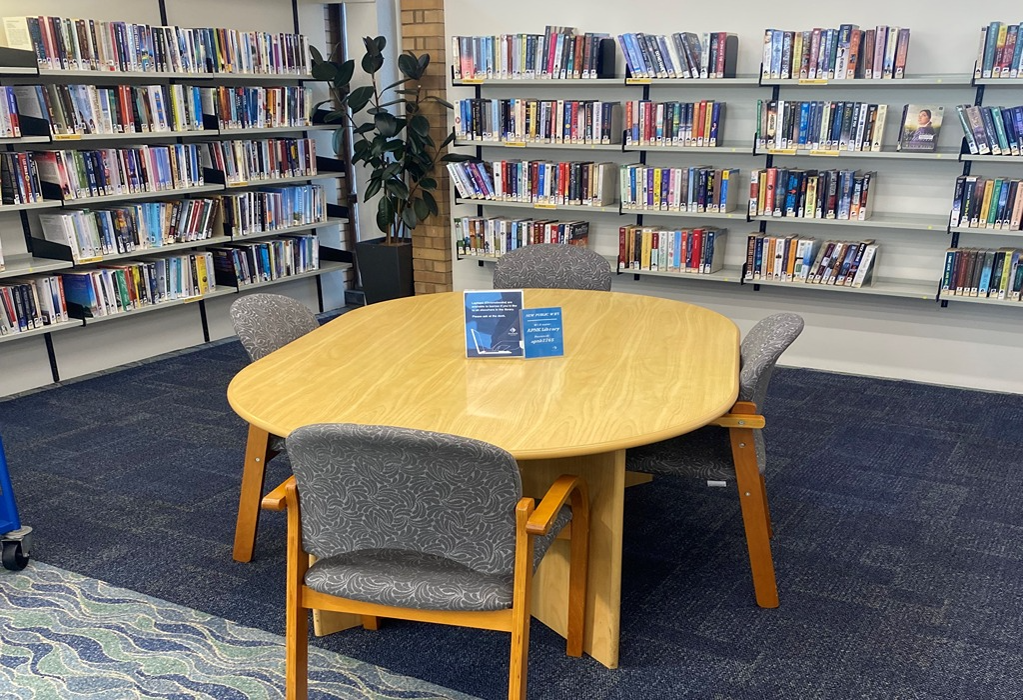 Round light timber table and four grey patterned armchairs on dark blue carpet. Wall-mounted grey metal bookshelves line the walls. Small blue signs are placed in the centre of the table.