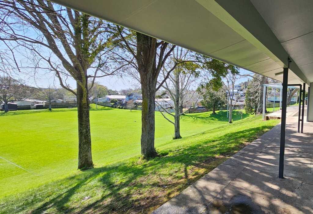 Level concrete walkway under a grey awning with dark poles. A grassy bank slopes down to a flat field. A concrete ramp with metal handrails is visible in the distance on the right.