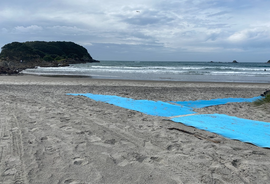 Bright blue textured accessibility mat laid over grey sand, creating a firm, level pathway towards the water. The matting consists of wide, overlapping sections for easier beach access.
