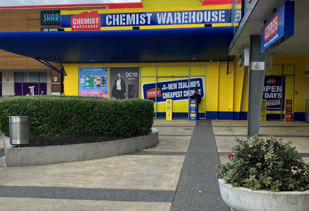 Wide, level concrete pathway leads to glass doors at a Chemist Warehouse. A large hedge in a planter and a bin are to the left. Bright yellow storefront with blue awning and clear signage.