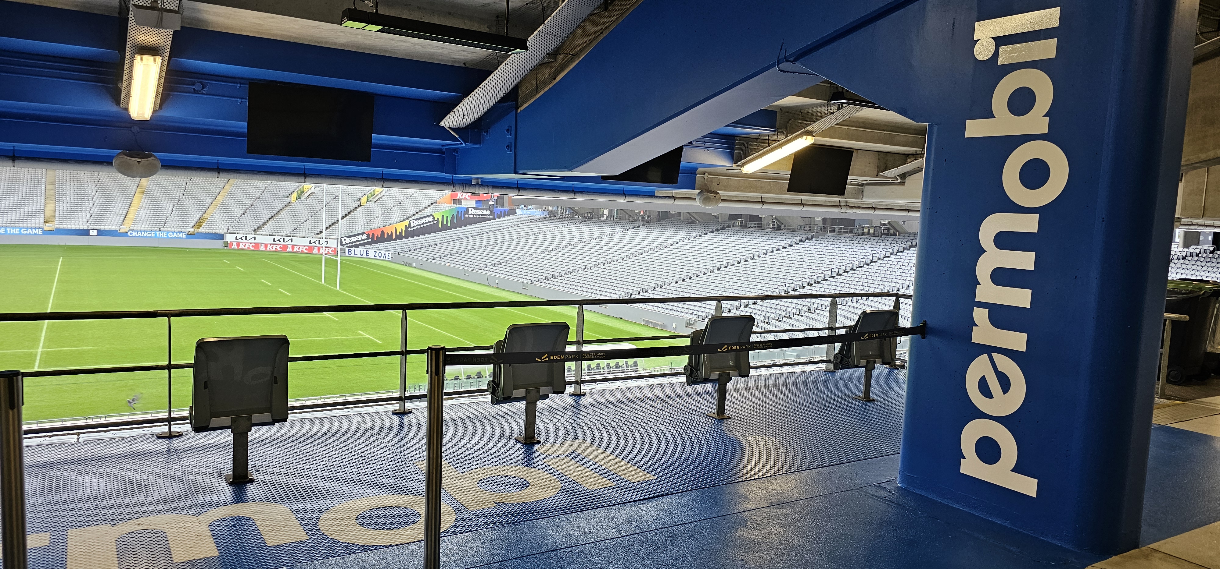 Level blue non-slip platform with four grey folding seats. Metal handrails and a retractable barrier overlook a green pitch and white stadium stands. Permobil logo on a large blue pillar.