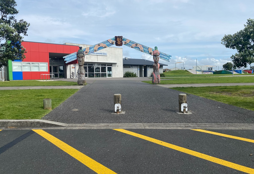 Level asphalt pathway leads from a car park with yellow lines to a community centre entrance. Two wooden bollards stand at the wide entry under a carved archway. Smooth level access throughout.