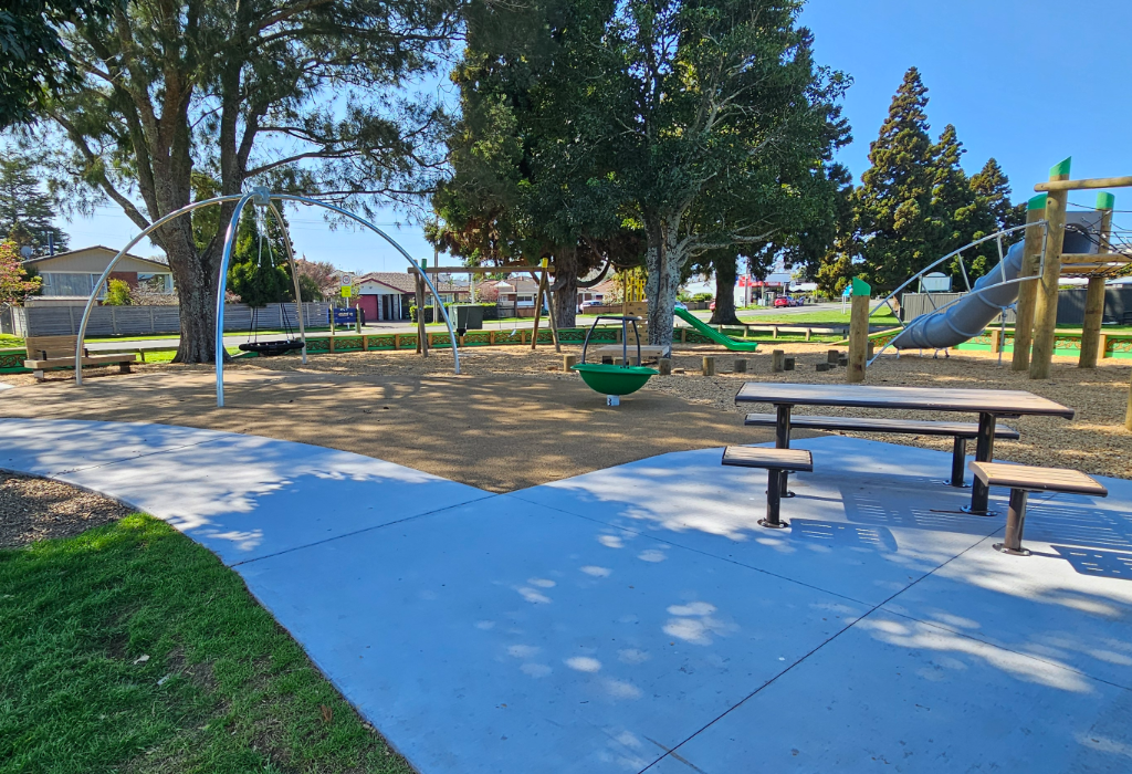 Level light blue concrete pathway leads to a timber picnic table. Tan soft-fall surfacing sits under playground equipment. Wooden bench seating is provided in the level, partially shaded area.