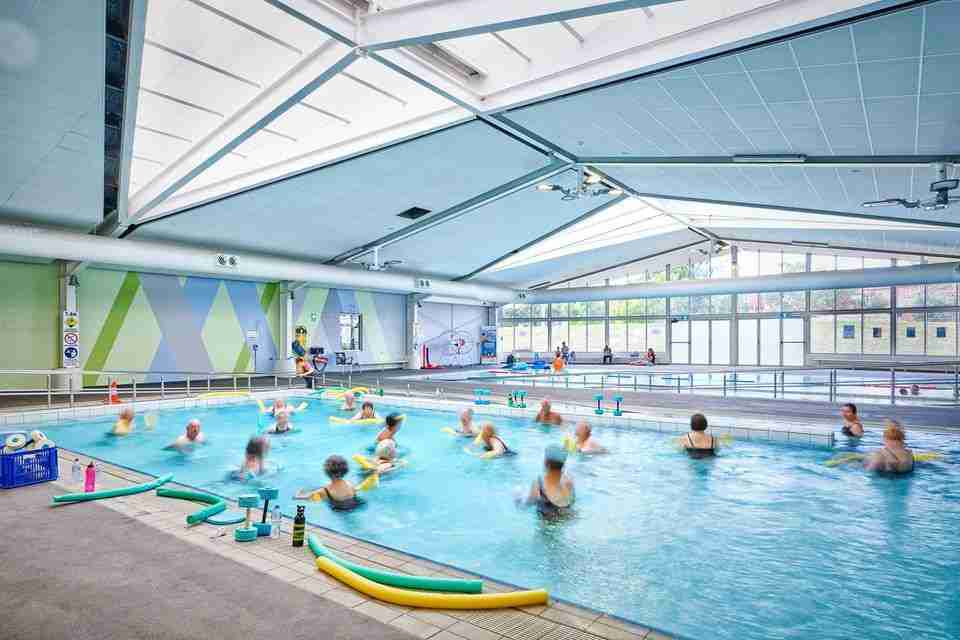 Level tiled indoor pool deck with perimeter handrails. Large glass windows and doors at far end. Brightly lit aquatic centre with high ceilings, wide pathways and ramp access.