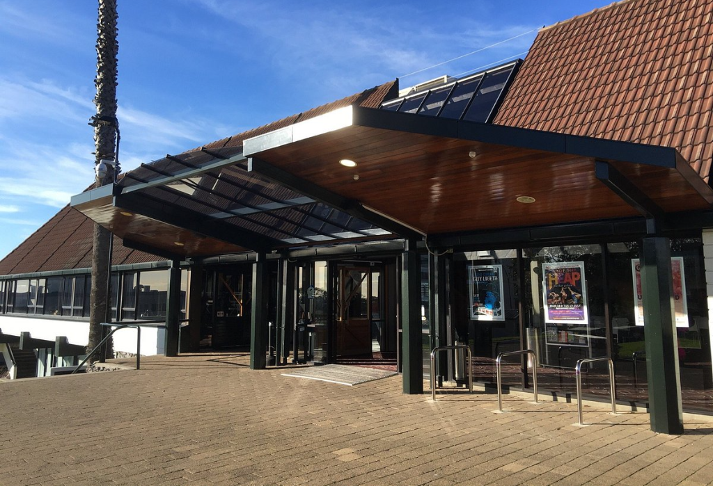 Level paved path leads to wide glass automatic doors under a large timber-lined canopy with recessed lights. A metal handrail is on the left and silver bike racks are on the right.