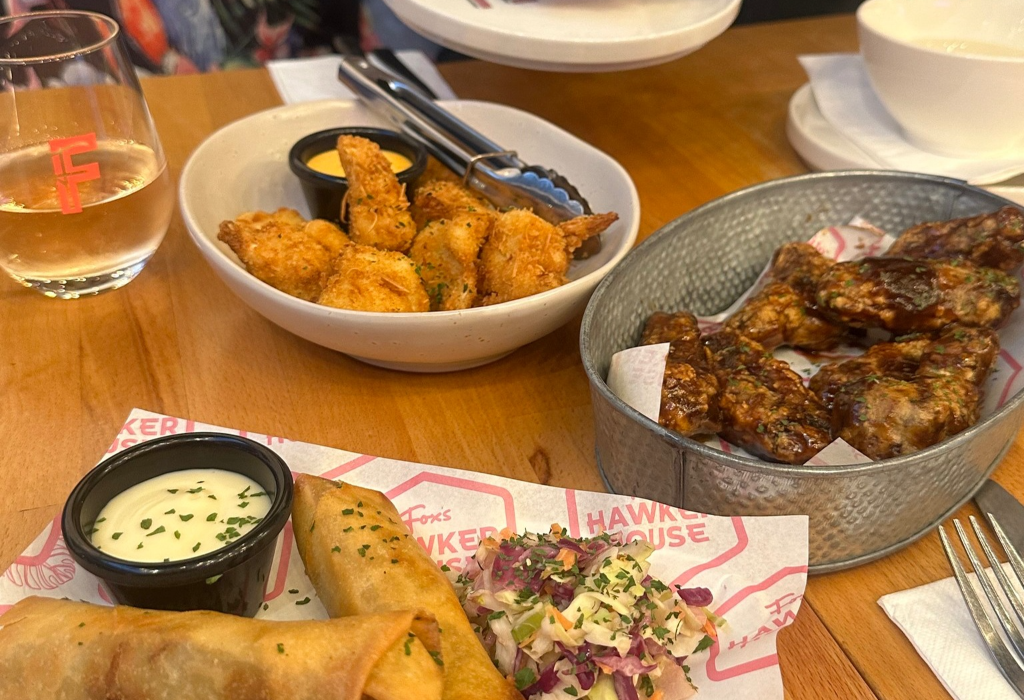 Wooden table featuring shared plates of fried spring rolls with slaw, battered food with dipping sauce, and glazed chicken wings in a metal bucket. A glass of pink liquid is on the left.