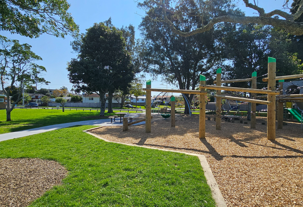 Level concrete pathway leads to picnic tables and a wooden playground structure on woodchip mulch. The area features flat mown grass and large leafy trees providing shade.