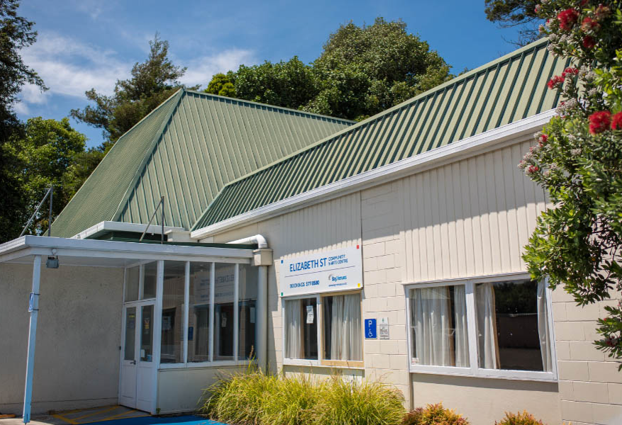 Level entrance to the Elizabeth St Community & Arts Centre. White glass doors under a porch. Blue accessible parking sign on wall and blue markings on the level concrete ground.