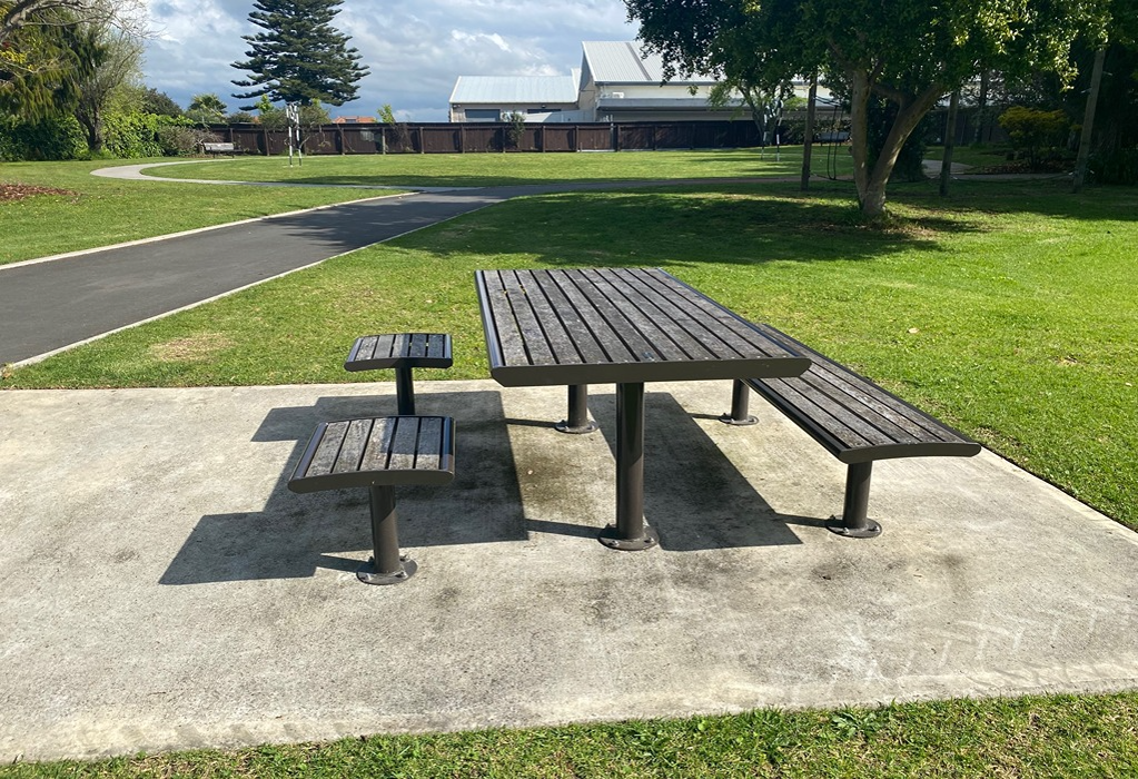 Picnic table on level concrete with a long wooden bench and two separate stools, creating wheelchair-accessible space. A smooth asphalt pathway borders the surrounding grassed park area.