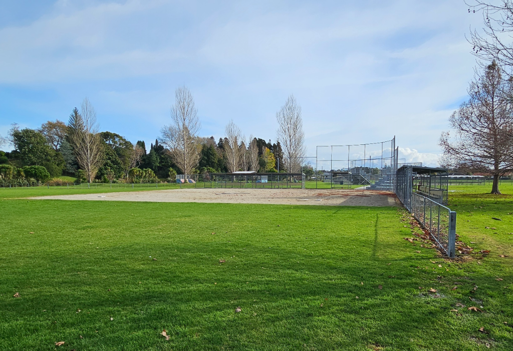 Level grass field leads to a flat dirt sports diamond. A chain-link fence encloses a dugout with bench seating under a metal roof. Terrain is flat with no visible paved pathways or ramps.